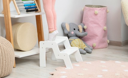 Photo of Little girl standing on step stool and reaching for toys on shelf indoors