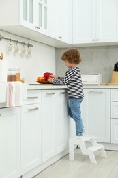 Photo of Little boy standing on step stool near countertop with fruits in kitchen