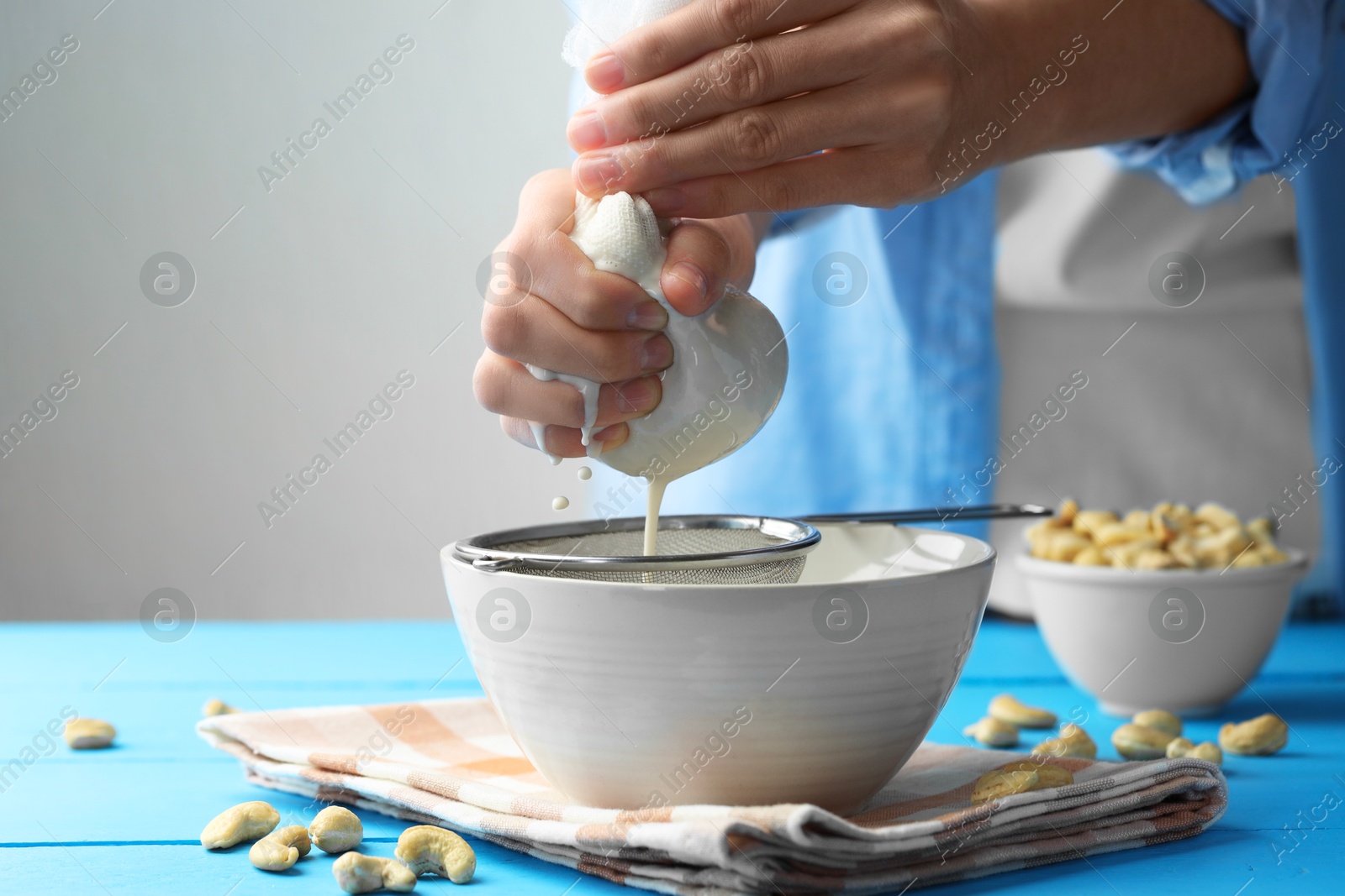 Photo of Woman straining cashew milk into bowl at light blue wooden table with nuts indoors, closeup