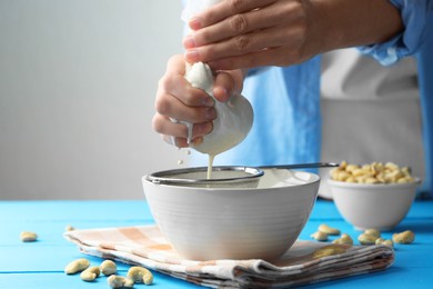 Photo of Woman straining cashew milk into bowl at light blue wooden table with nuts indoors, closeup