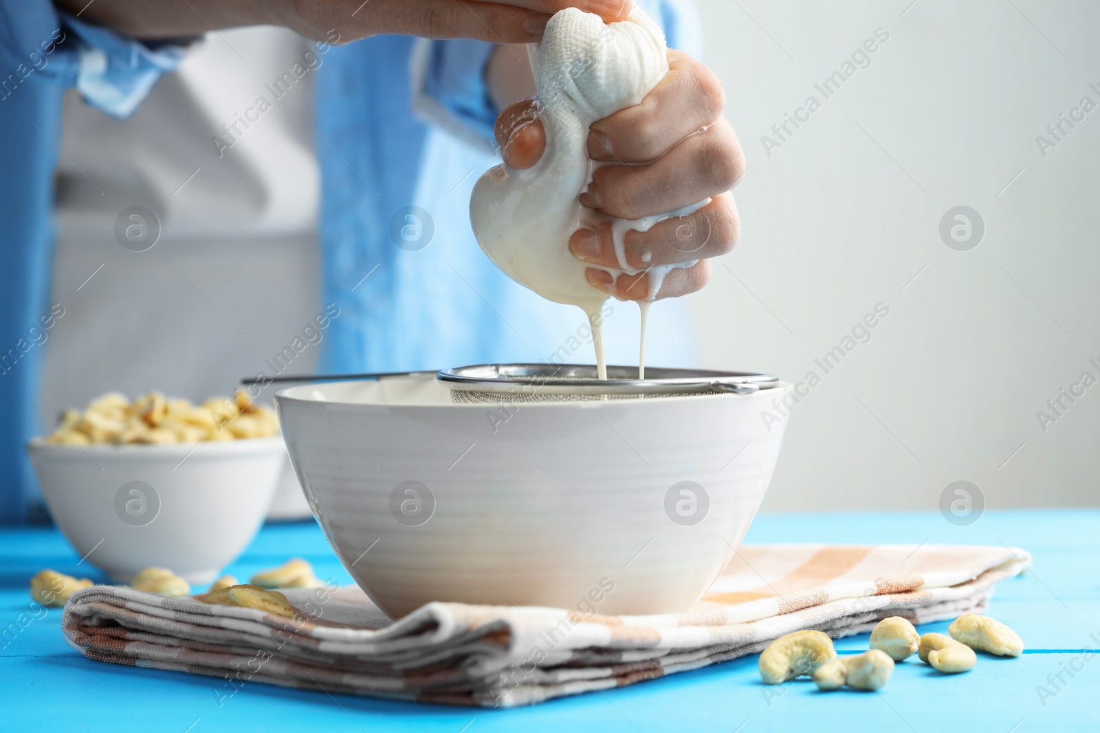 Photo of Woman straining cashew milk into bowl at light blue wooden table with nuts indoors, closeup