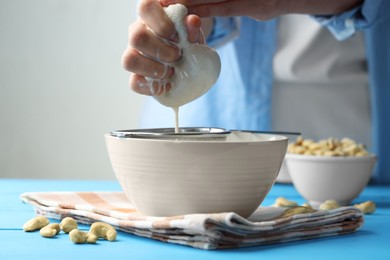 Photo of Woman straining cashew milk into bowl at light blue wooden table with nuts indoors, closeup