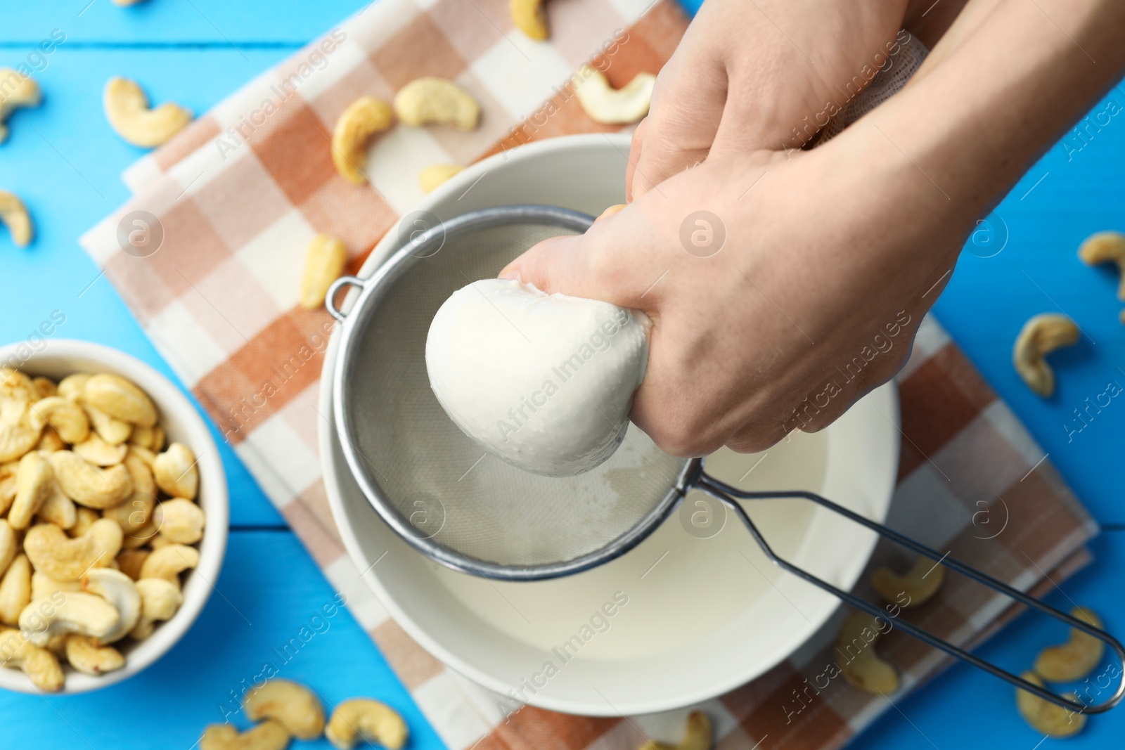 Photo of Woman straining cashew milk into sieve and bowl at light blue wooden table with nuts, top view