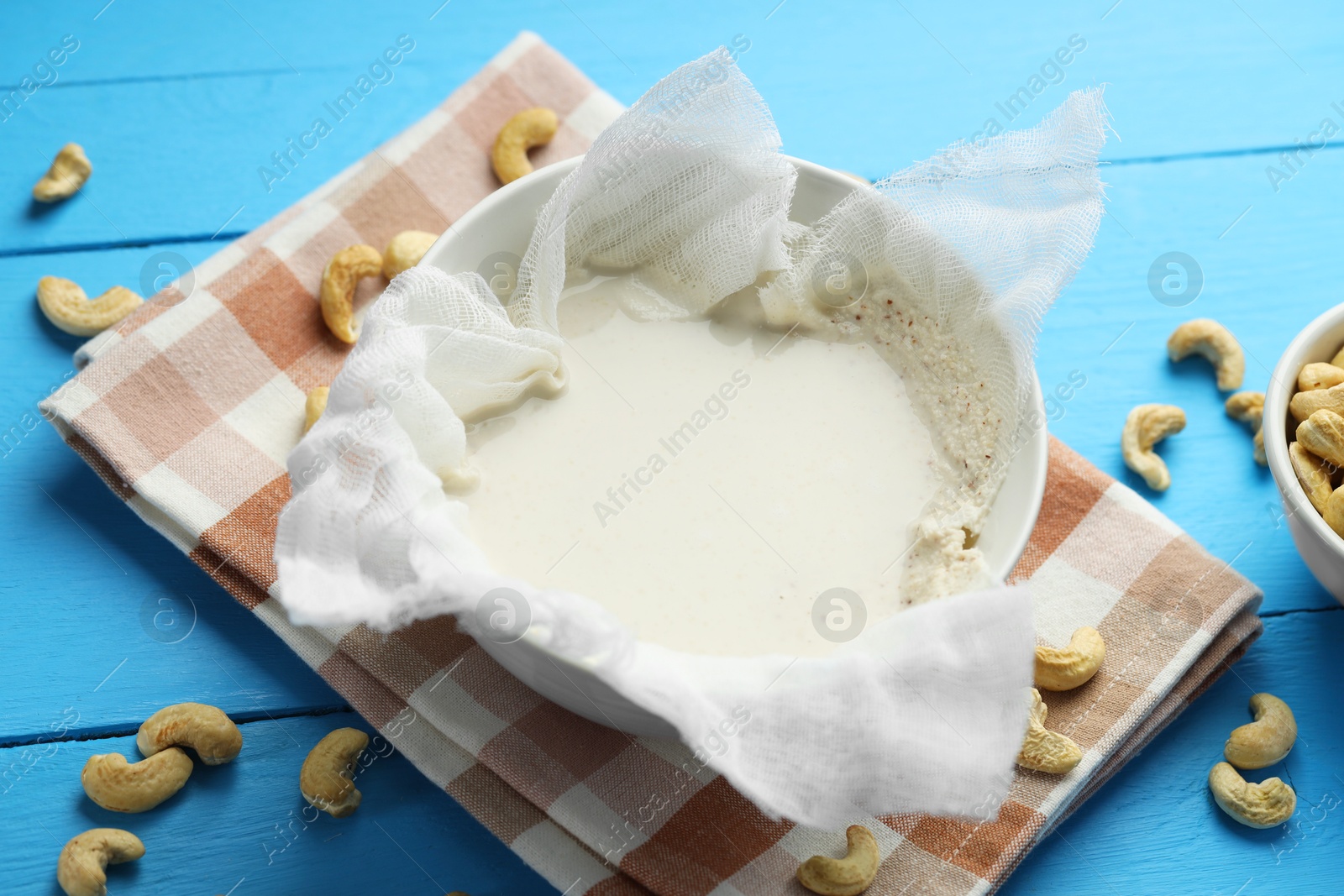 Photo of Making cashew milk. Cheesecloth with milk in bowl among nuts on light blue wooden table, closeup