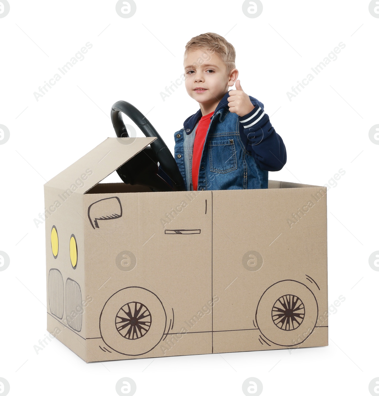 Photo of Little boy showing thumbs up while driving car made of cardboard on white background