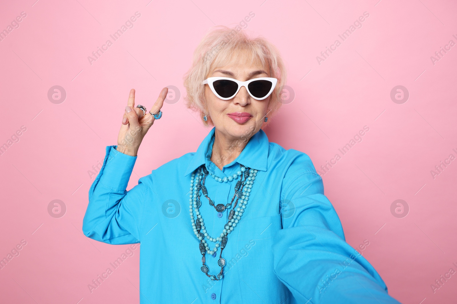 Photo of Cool grandmother showing rock gesture while taking selfie on pink background