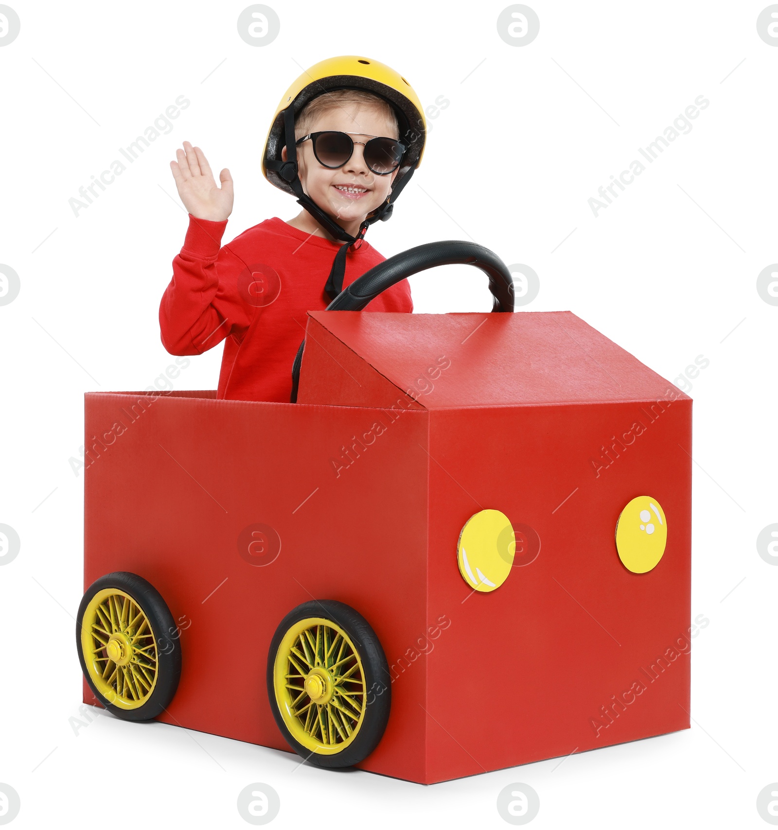 Photo of Little boy waving while driving car made of cardboard on white background