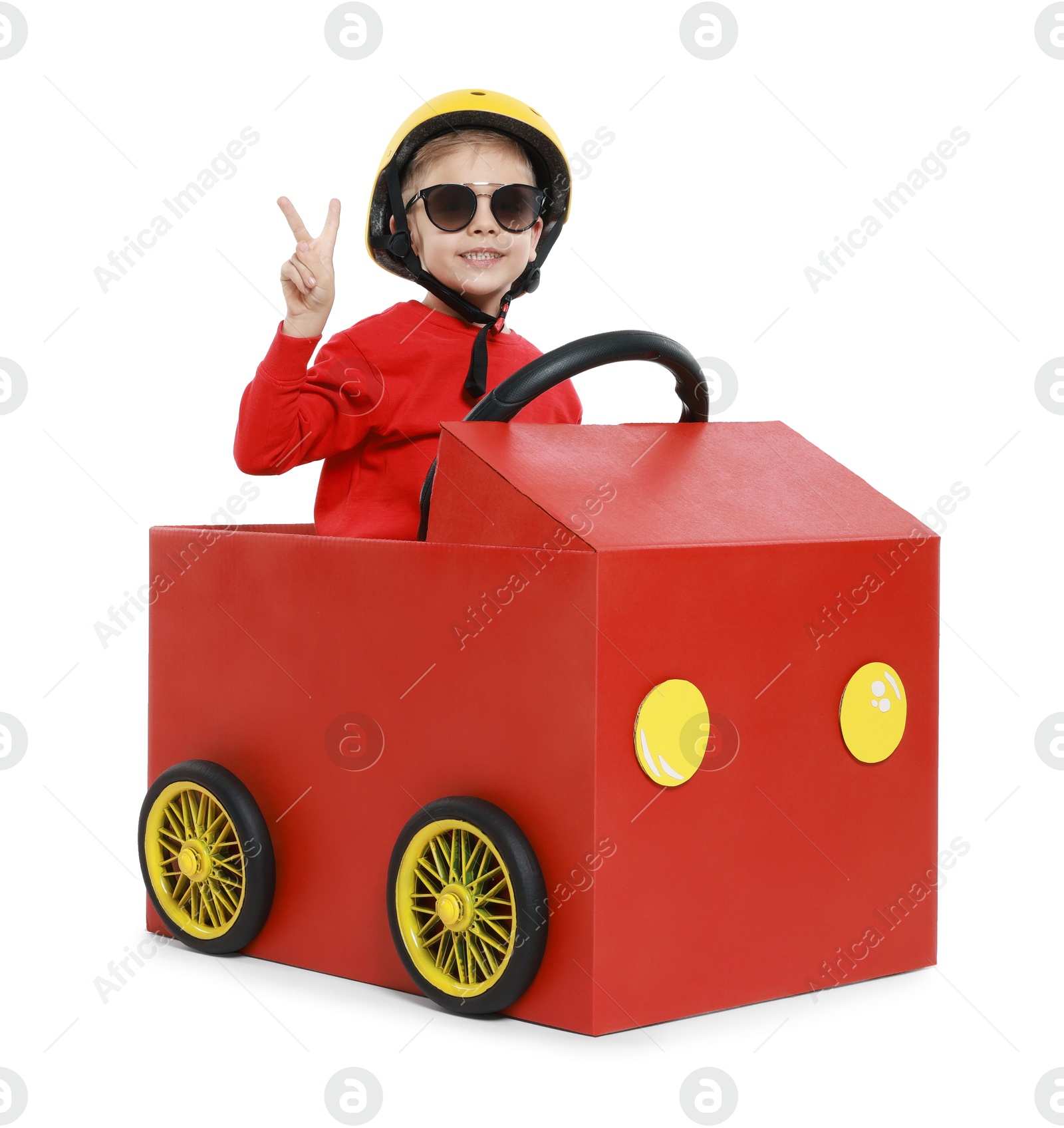 Photo of Little boy showing V-sign while driving car made of cardboard on white background