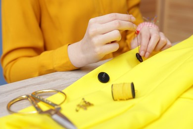 Photo of Woman sewing button onto jacket with thread at light wooden table indoors, closeup