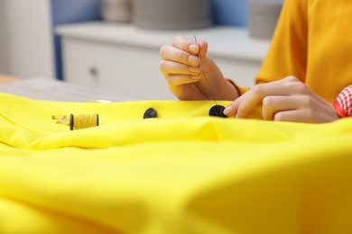 Photo of Woman sewing button onto jacket with thread at table indoors, closeup