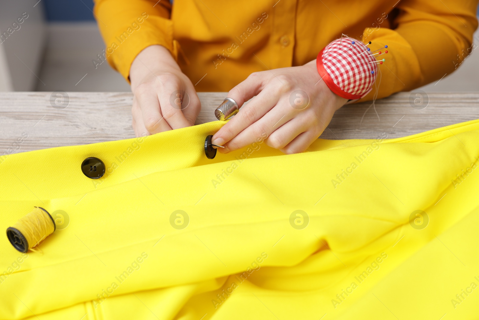 Photo of Woman sewing button onto jacket with thread at light wooden table indoors, closeup