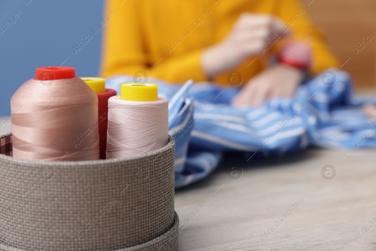 Photo of Woman working with cloth at light wooden table indoors, focus on spools of sewing threads