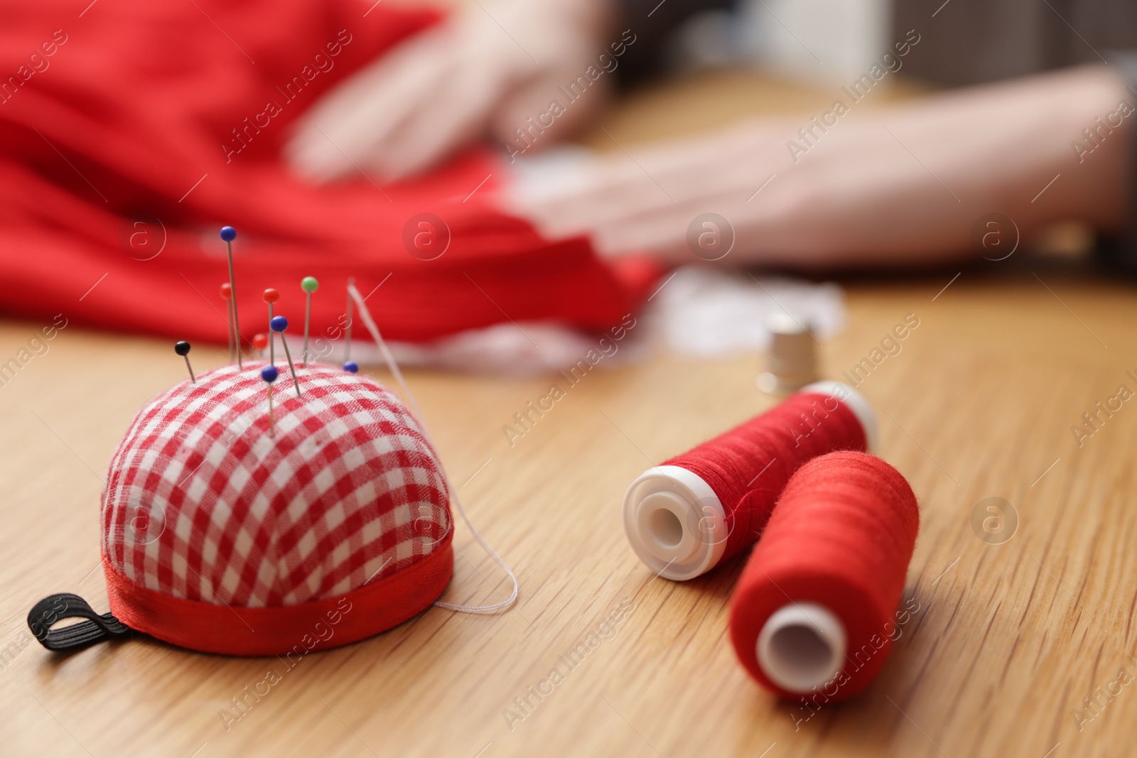 Photo of Woman working with cloth at wooden table, focus on spools of sewing threads and pincushion