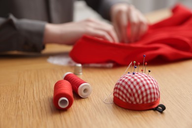 Photo of Woman working with cloth at wooden table, focus on spools of sewing threads and pincushion