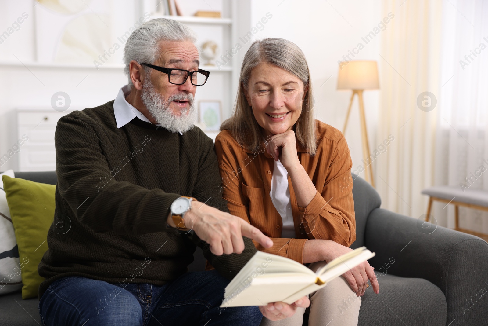 Photo of Cute elderly couple reading book together on sofa at home