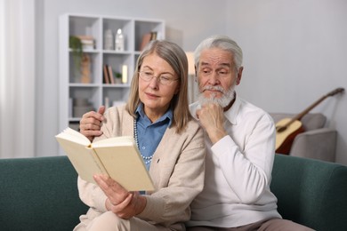 Photo of Cute elderly couple reading book together on sofa at home