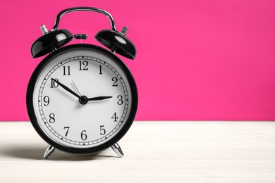 Photo of One black alarm clock on white wooden table against bright pink background, space for text