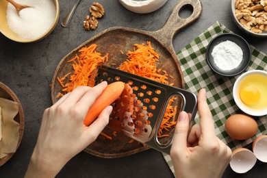 Photo of Making carrot cake. Woman grating vegetable at dark textured table with ingredients, top view