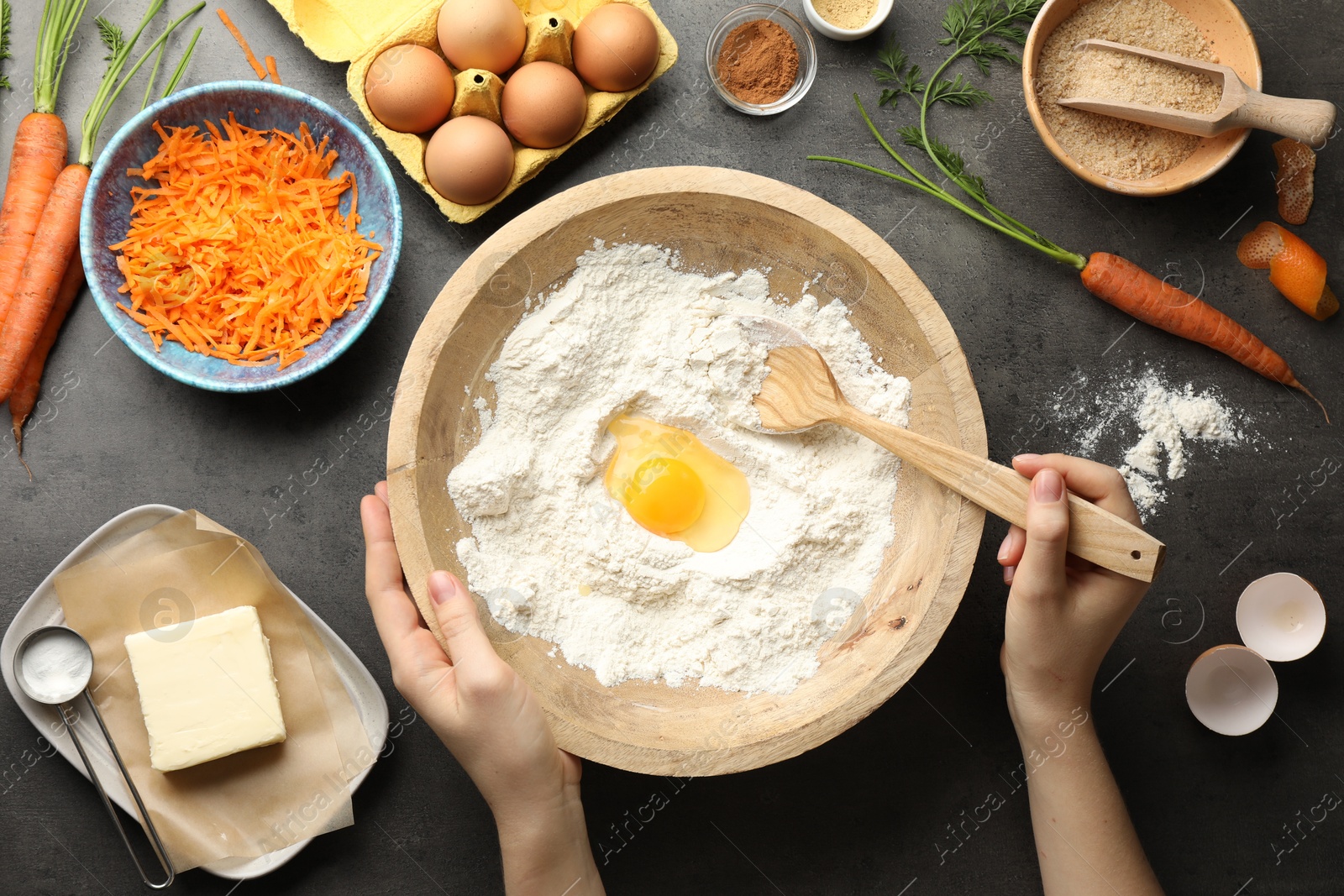 Photo of Woman making dough for carrot cake at dark textured table with ingredients, top view