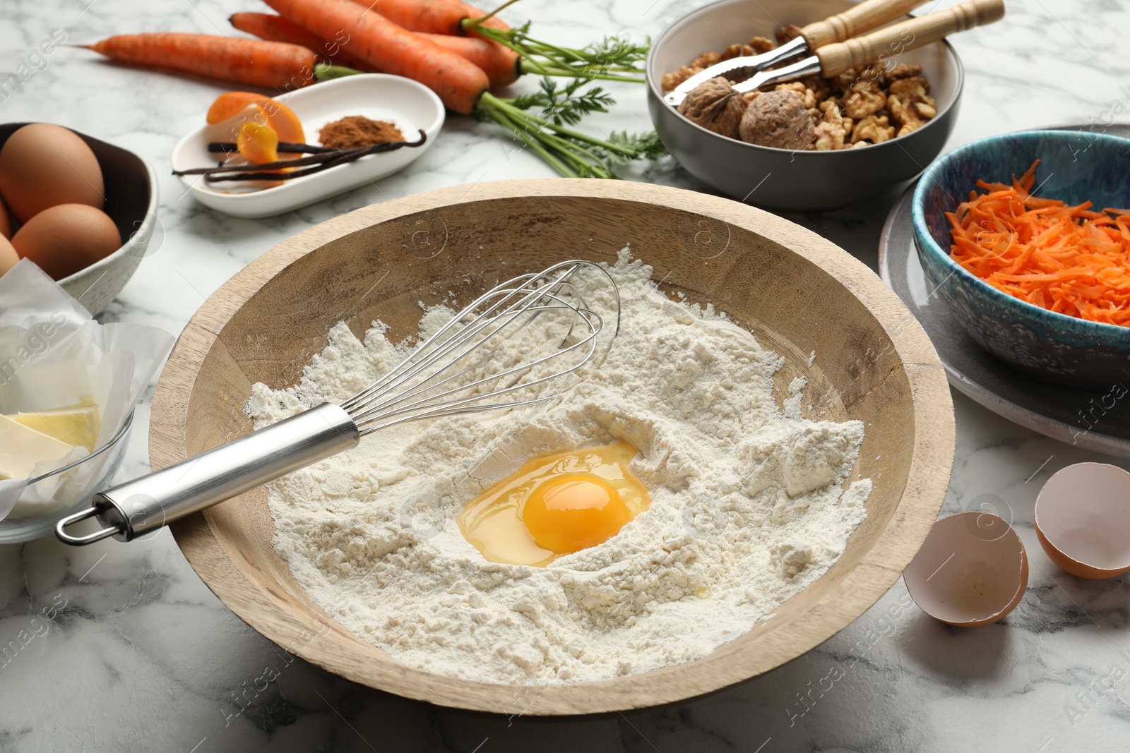 Photo of Making carrot cake. Bowl with flour, egg, whisk and other ingredients on white marble table, closeup