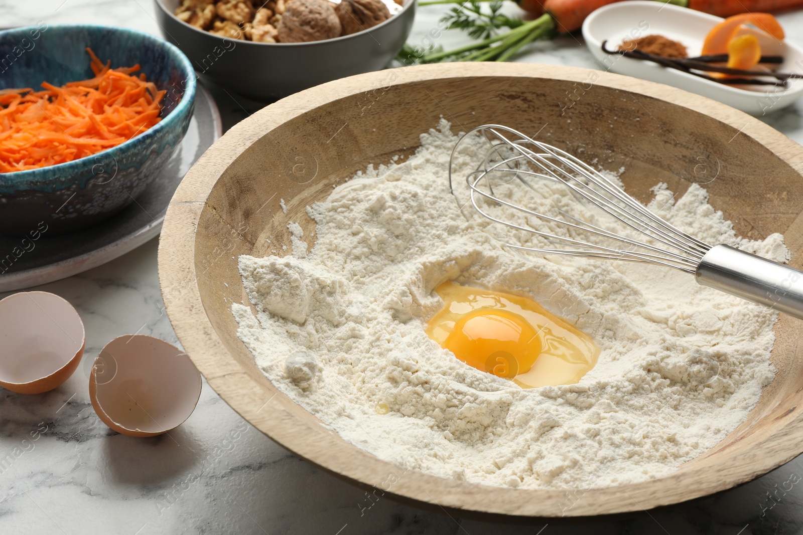 Photo of Making carrot cake. Bowl with flour, egg, whisk and other ingredients on white marble table, closeup