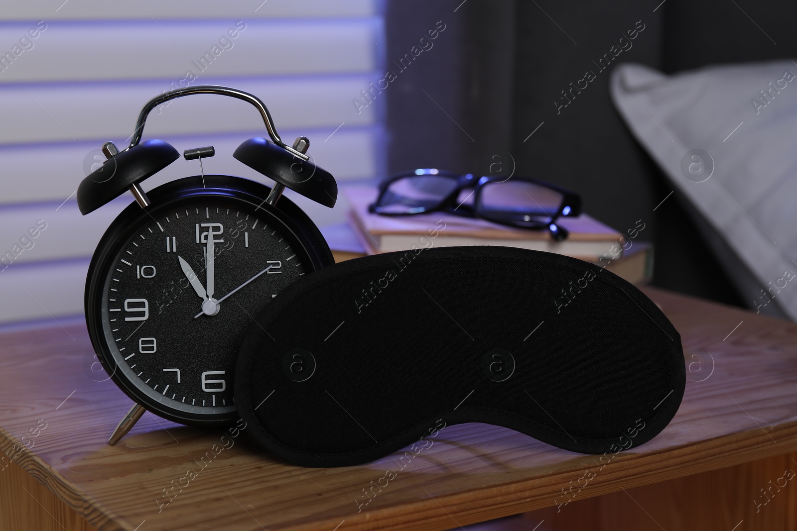 Photo of Alarm clock, sleeping mask, books and glasses on nightstand near bed