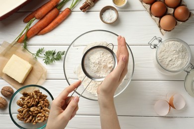 Photo of Making carrot cake. Woman sifting flour into bowl at white wooden table, top view