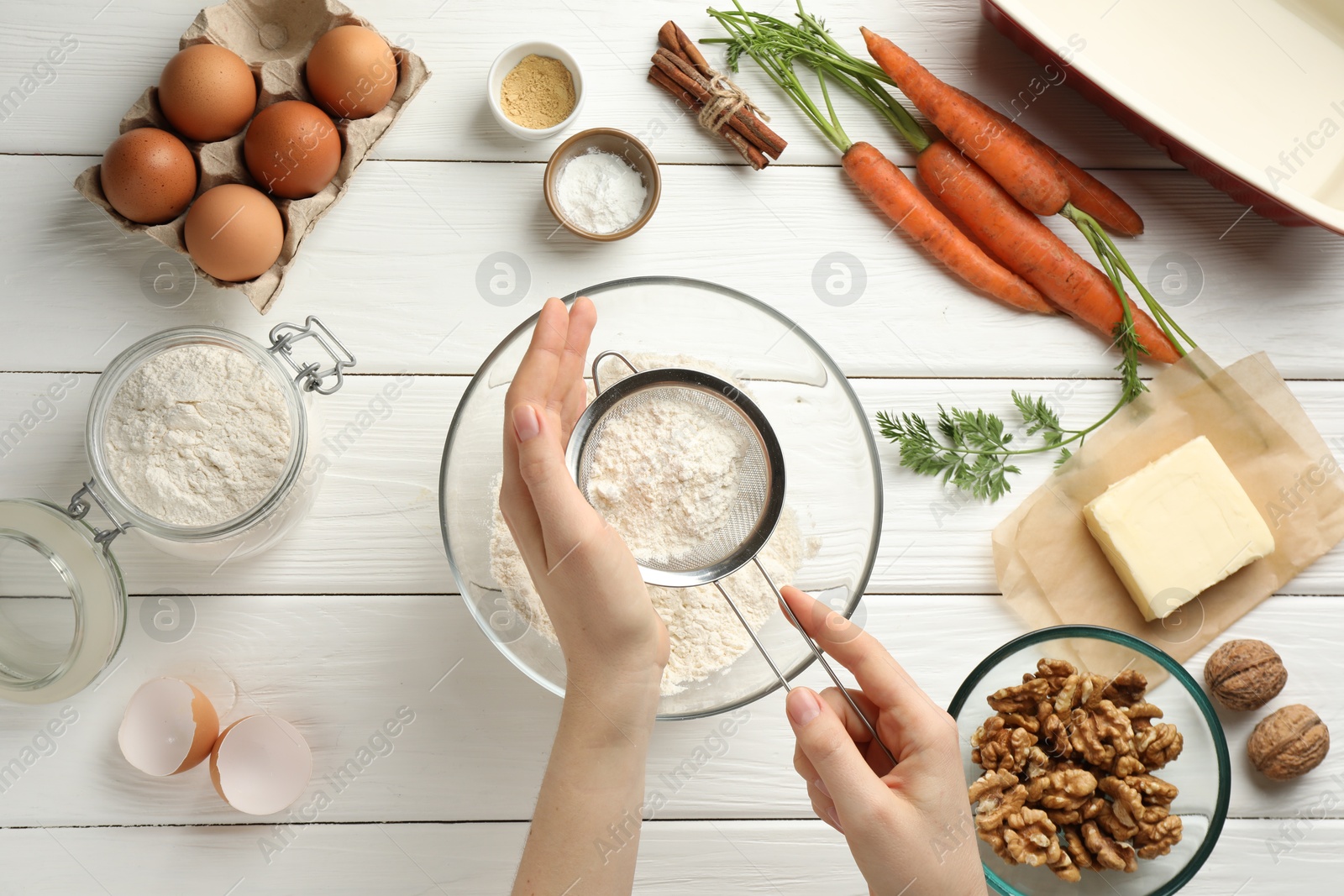 Photo of Making carrot cake. Woman sifting flour into bowl at white wooden table, top view