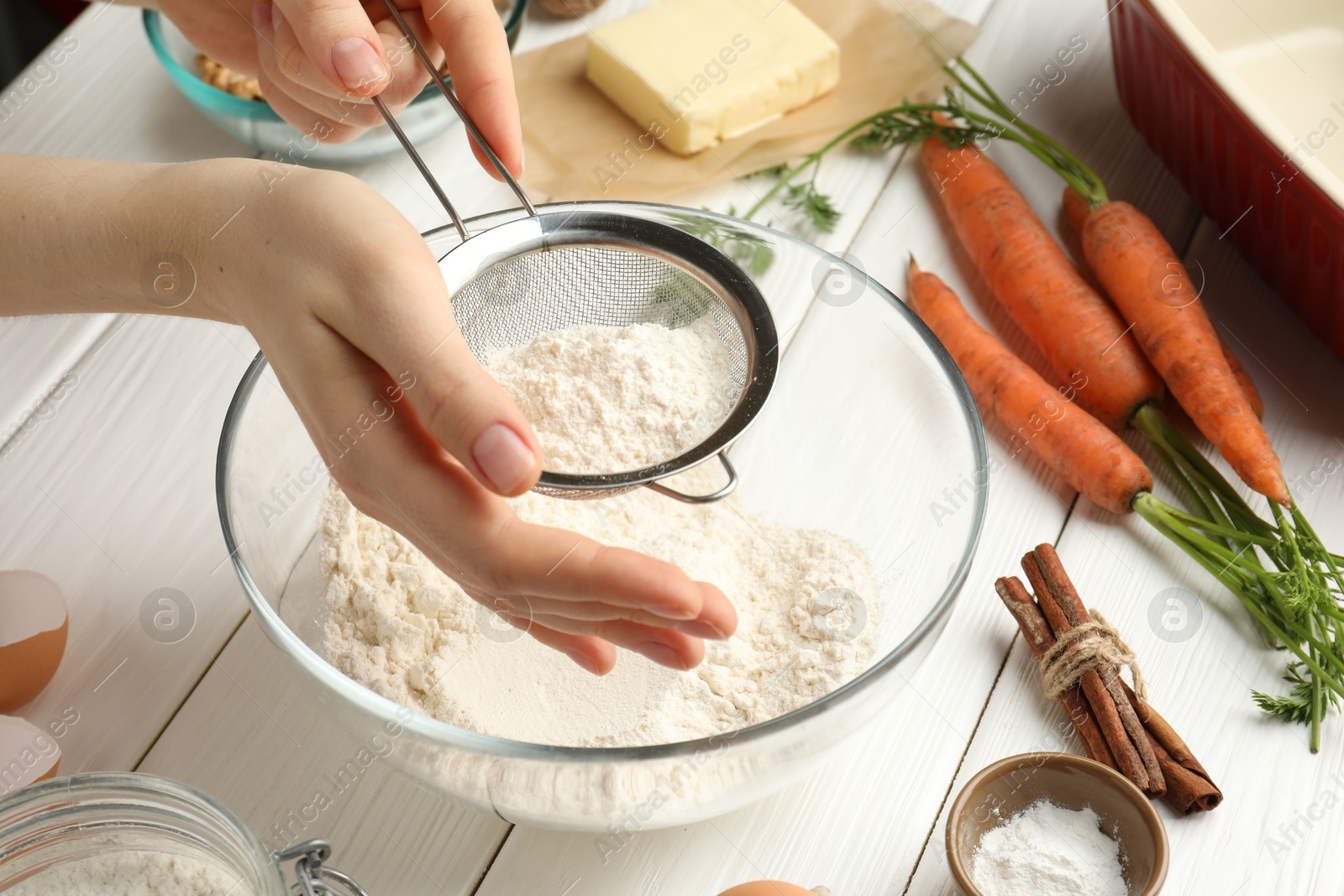 Photo of Making carrot cake. Woman sifting flour into bowl at white wooden table, closeup