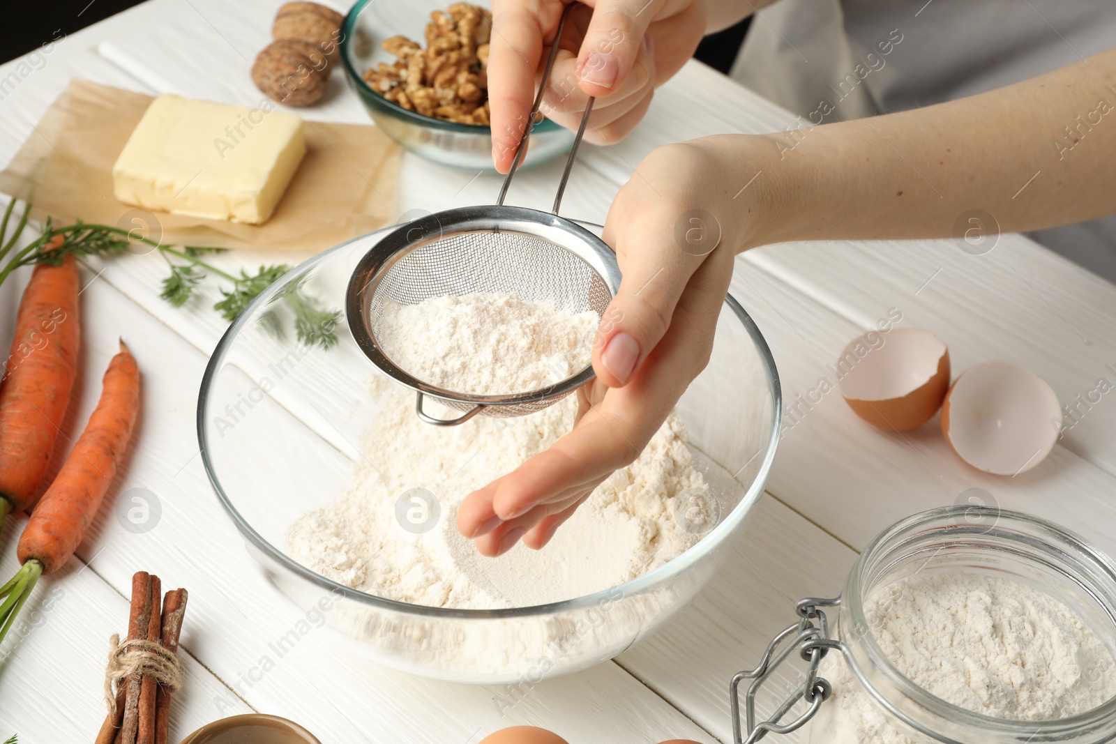 Photo of Making carrot cake. Woman sifting flour into bowl at white wooden table, closeup