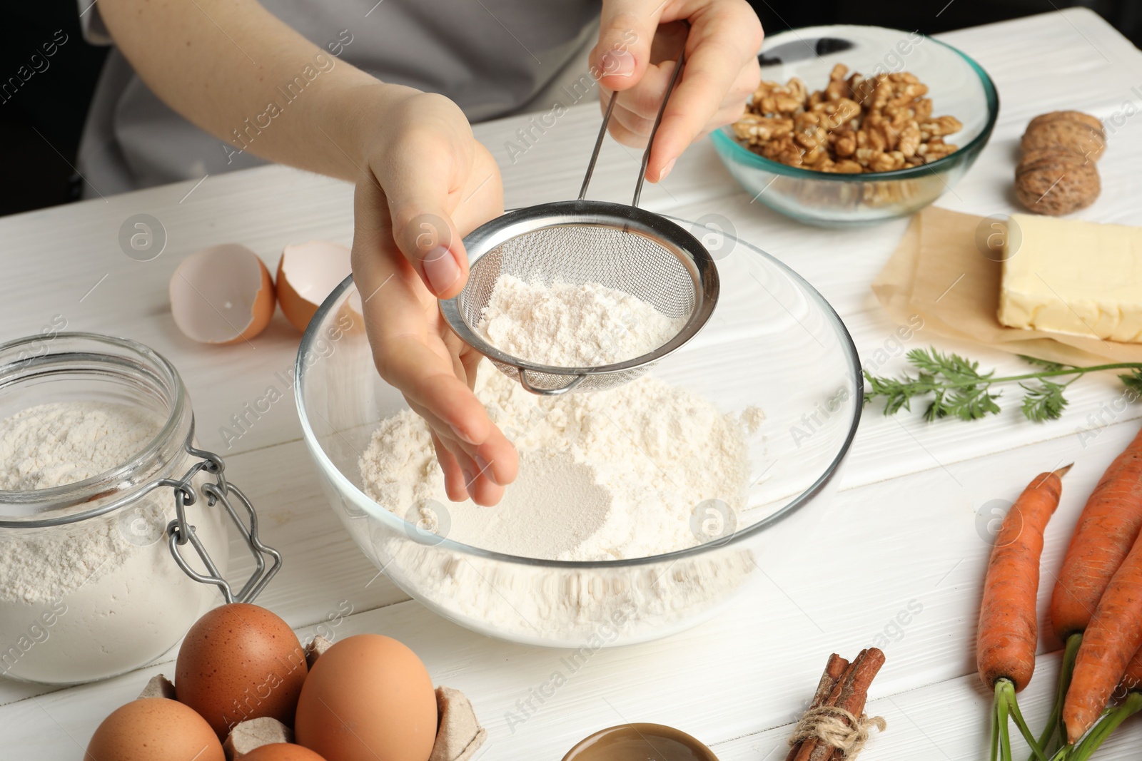 Photo of Making carrot cake. Woman sifting flour into bowl at white wooden table, closeup