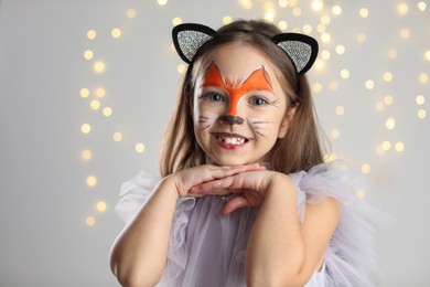 Photo of Cute girl with painted face and ears as cat on grey background with blurred lights, space for text