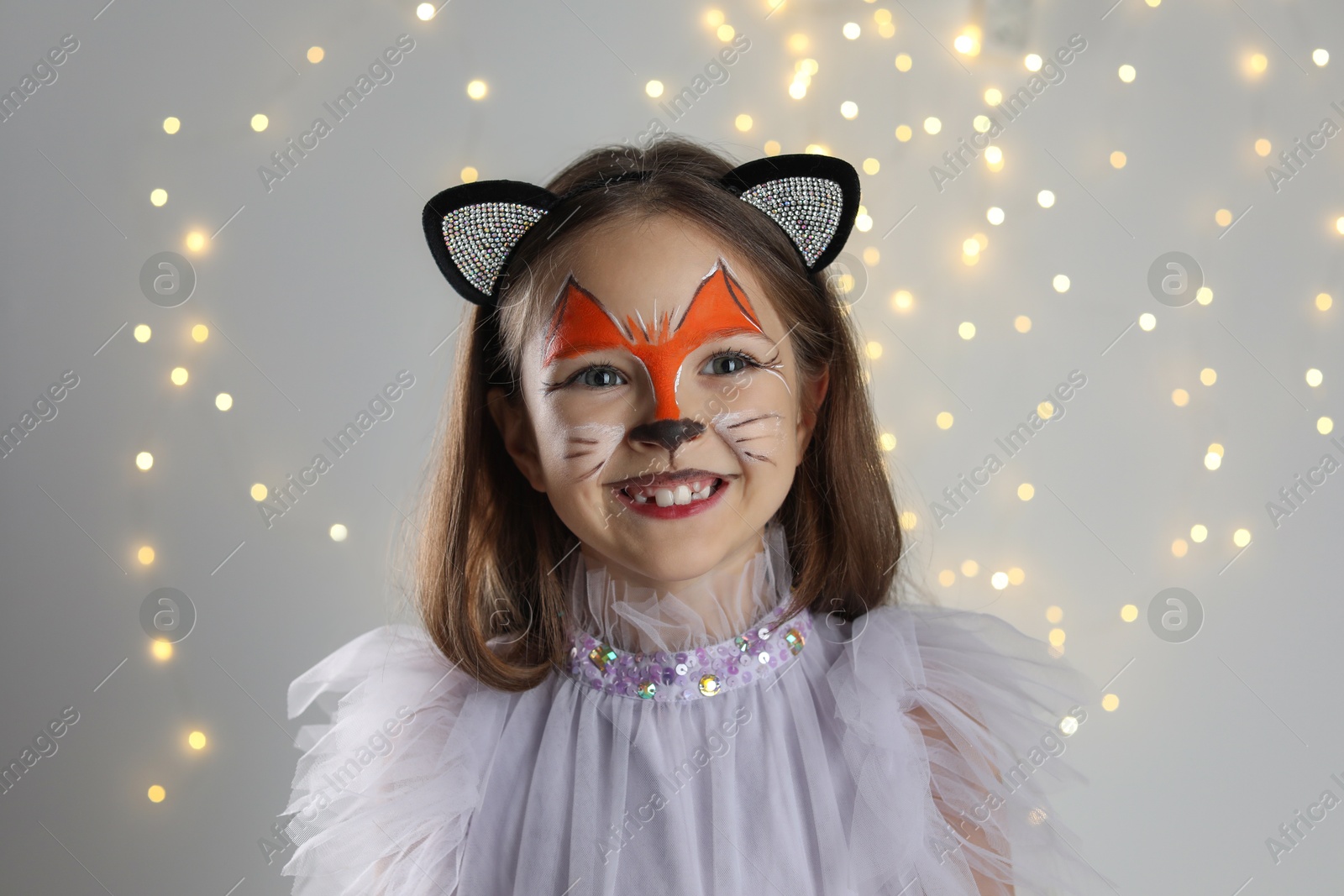 Photo of Cute girl with painted face and ears as cat on grey background with blurred lights