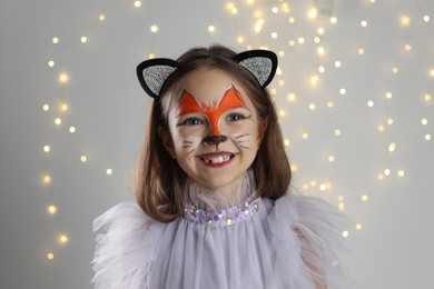 Cute girl with painted face and ears as cat on grey background with blurred lights