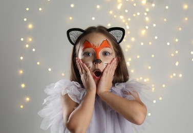 Photo of Cute girl with painted face and ears as cat on grey background with blurred lights