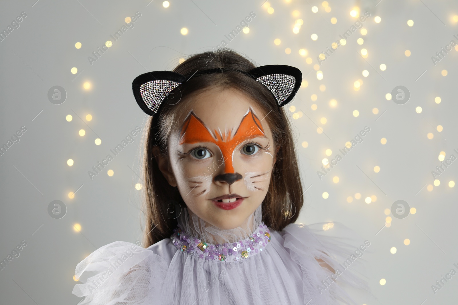 Photo of Cute girl with painted face and ears as cat on grey background with blurred lights