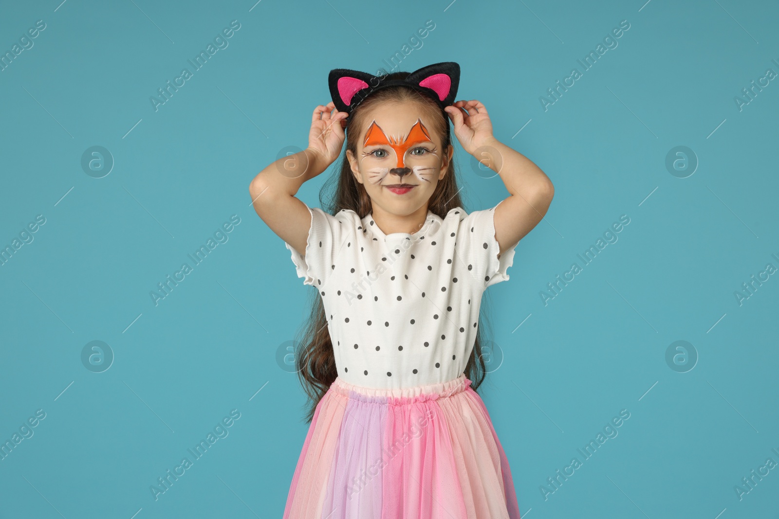 Photo of Cute girl with painted face and ears as cat on light blue background