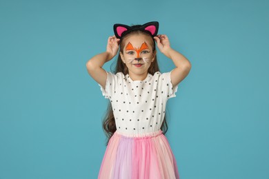 Cute girl with painted face and ears as cat on light blue background