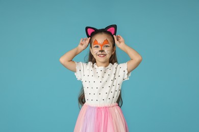 Photo of Cute girl with painted face and ears as cat on light blue background