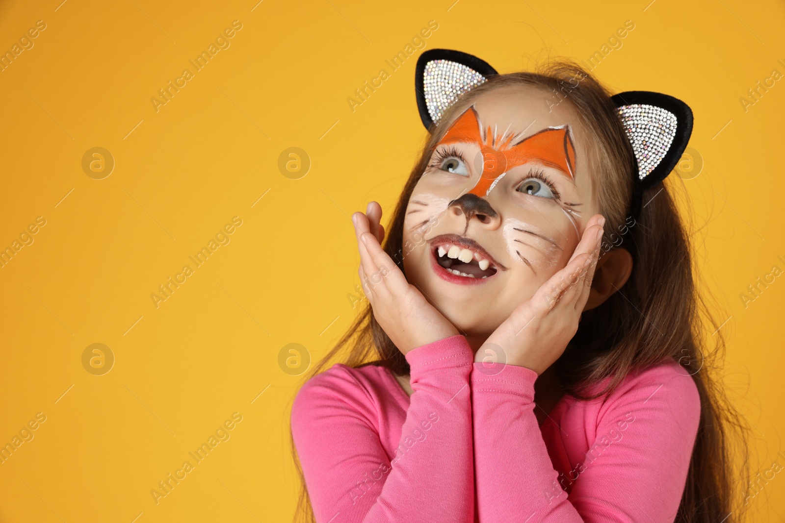 Photo of Cute girl with painted face and ears as cat on orange background, space for text