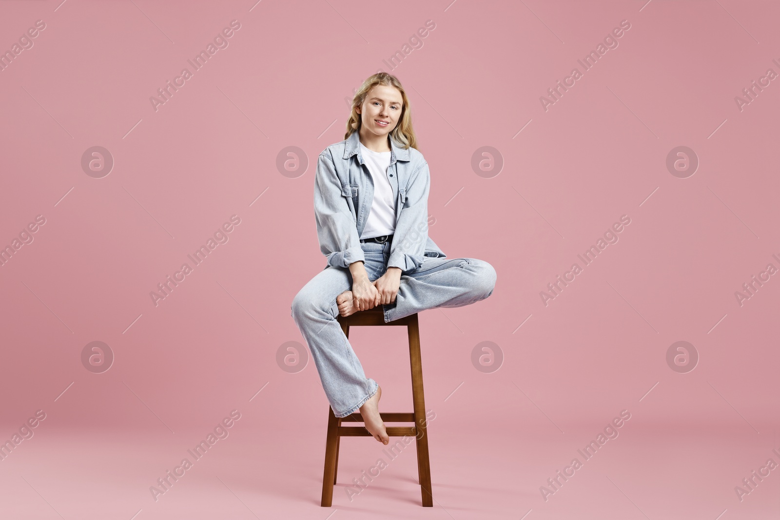 Photo of Smiling woman in stylish jeans sitting on stool against pink background