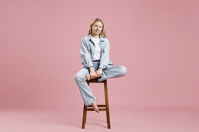 Smiling woman in stylish jeans sitting on stool against pink background