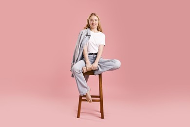 Smiling woman in stylish jeans sitting on stool against pink background