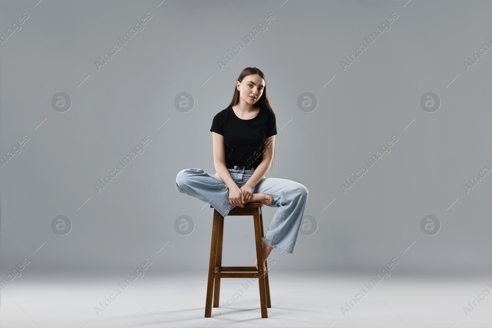 Photo of Beautiful young woman in stylish jeans sitting on stool against grey background