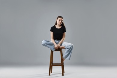 Beautiful young woman in stylish jeans sitting on stool against grey background