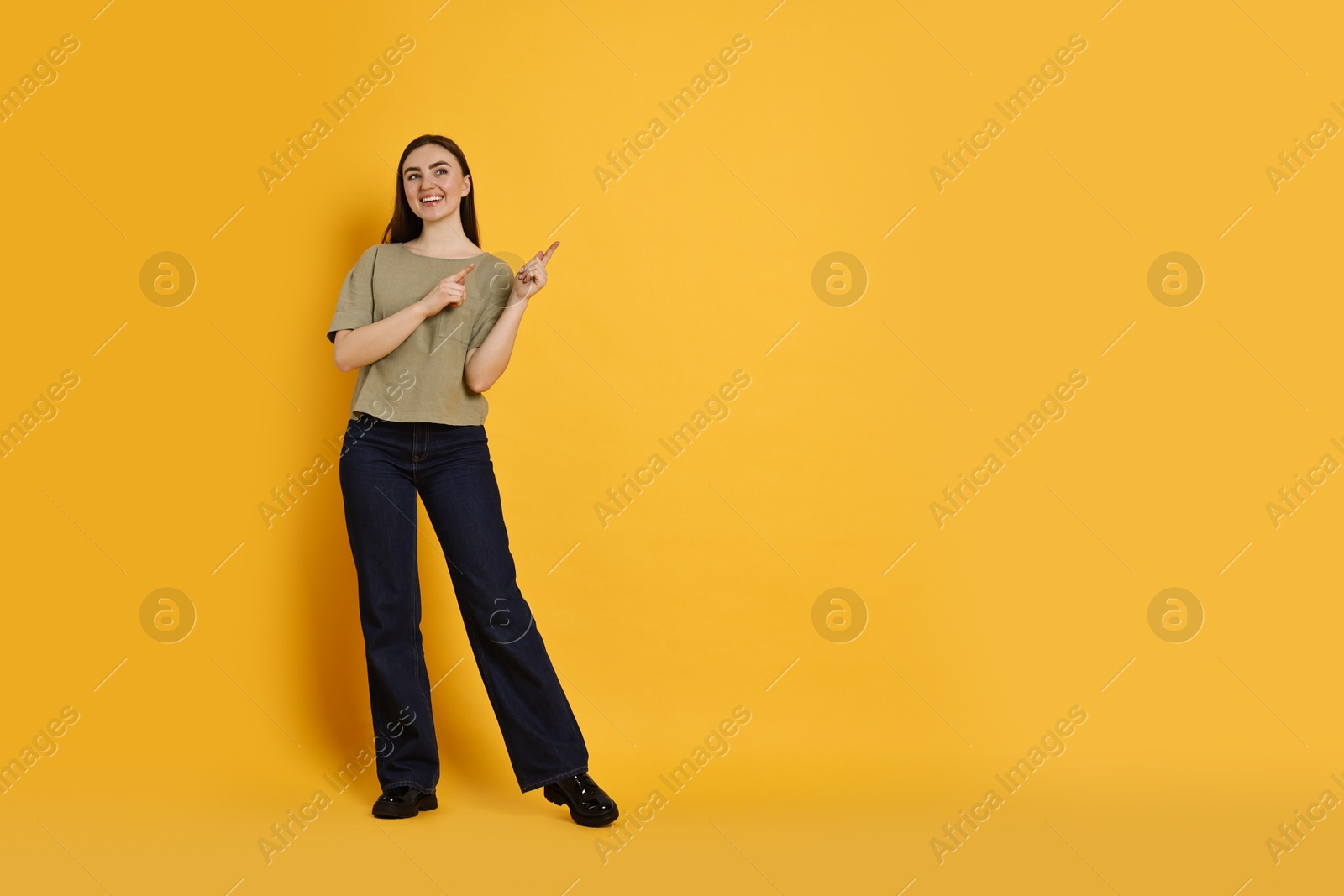 Photo of Smiling woman in stylish jeans pointing at something on orange background. Space for text