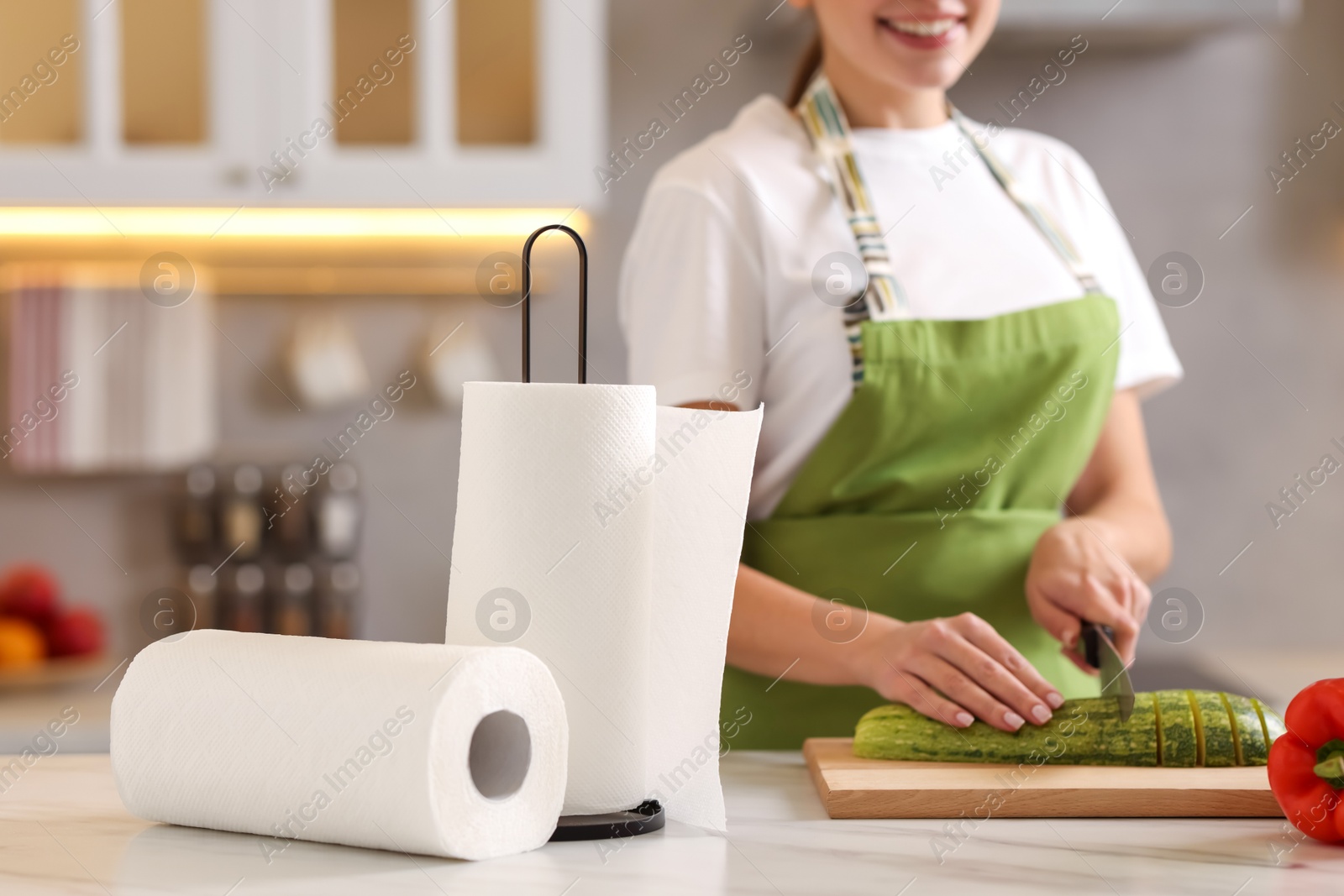 Photo of Woman cutting zucchini at white marble table in kitchen, focus on rolls of paper towels