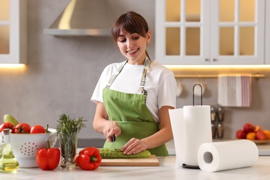 Photo of Woman cutting zucchini at white marble table with rolls of paper towels in kitchen