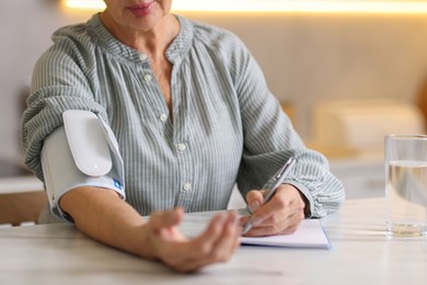 Photo of Senior woman writing results of blood pressure measurement in kitchen at home, closeup