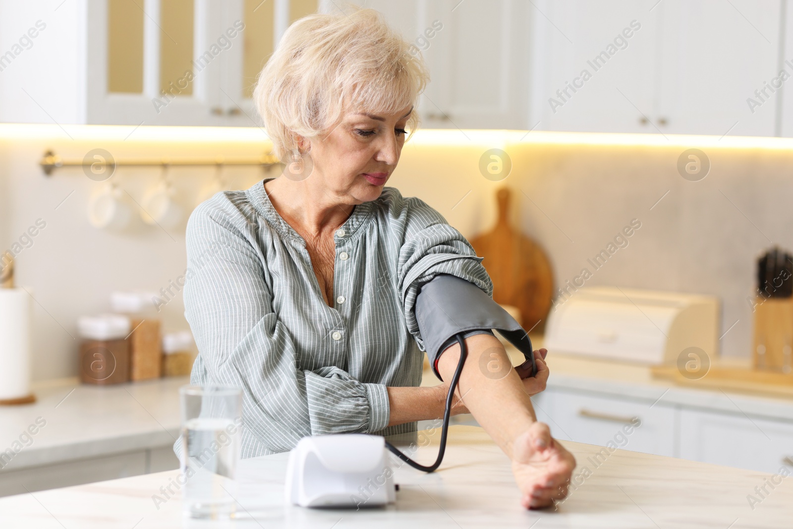 Photo of Senior woman measuring blood pressure at table in kitchen